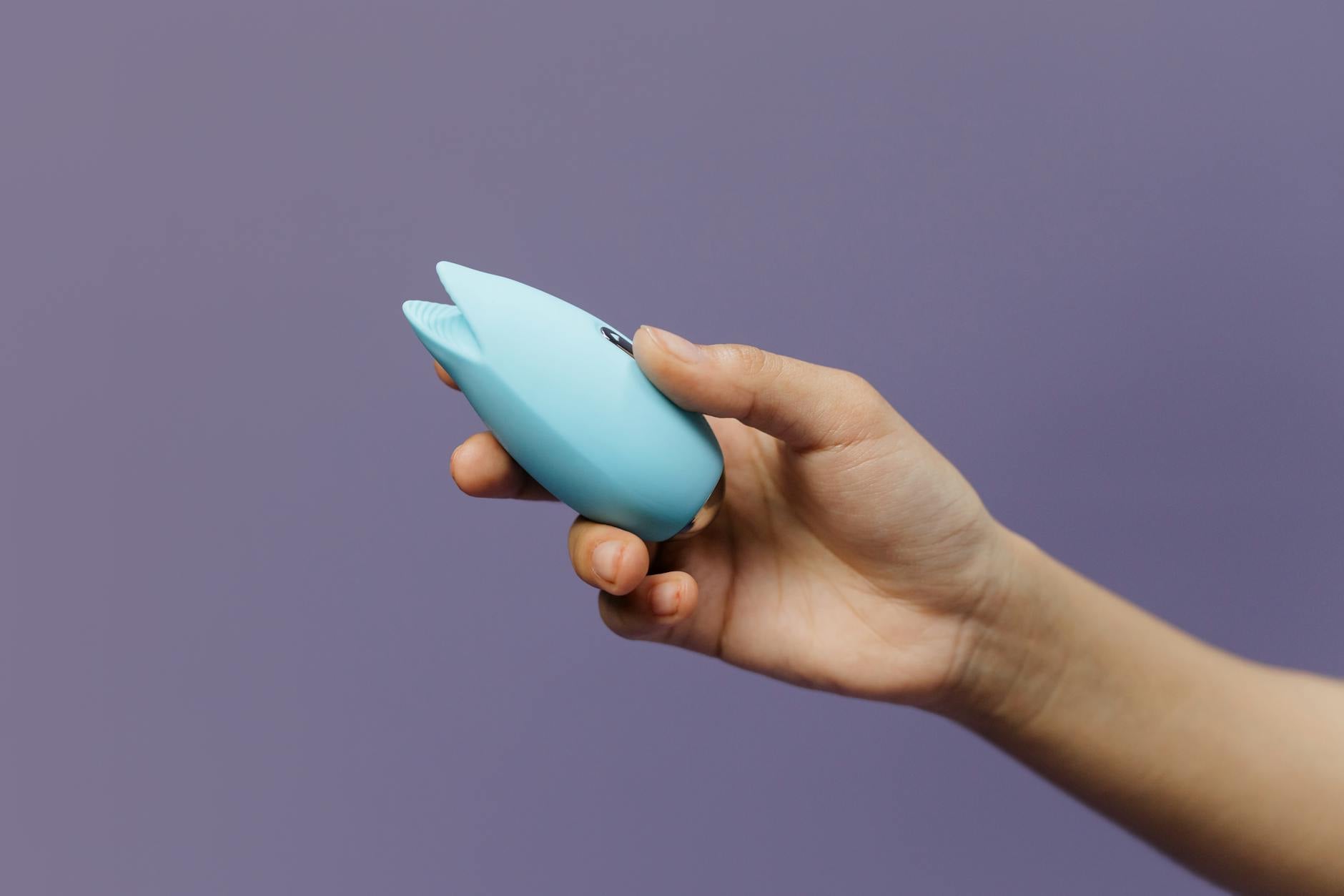 A blue silicone clitoral vibrator held in hand against a solid purple background, representing self-pleasure and personal intimacy.