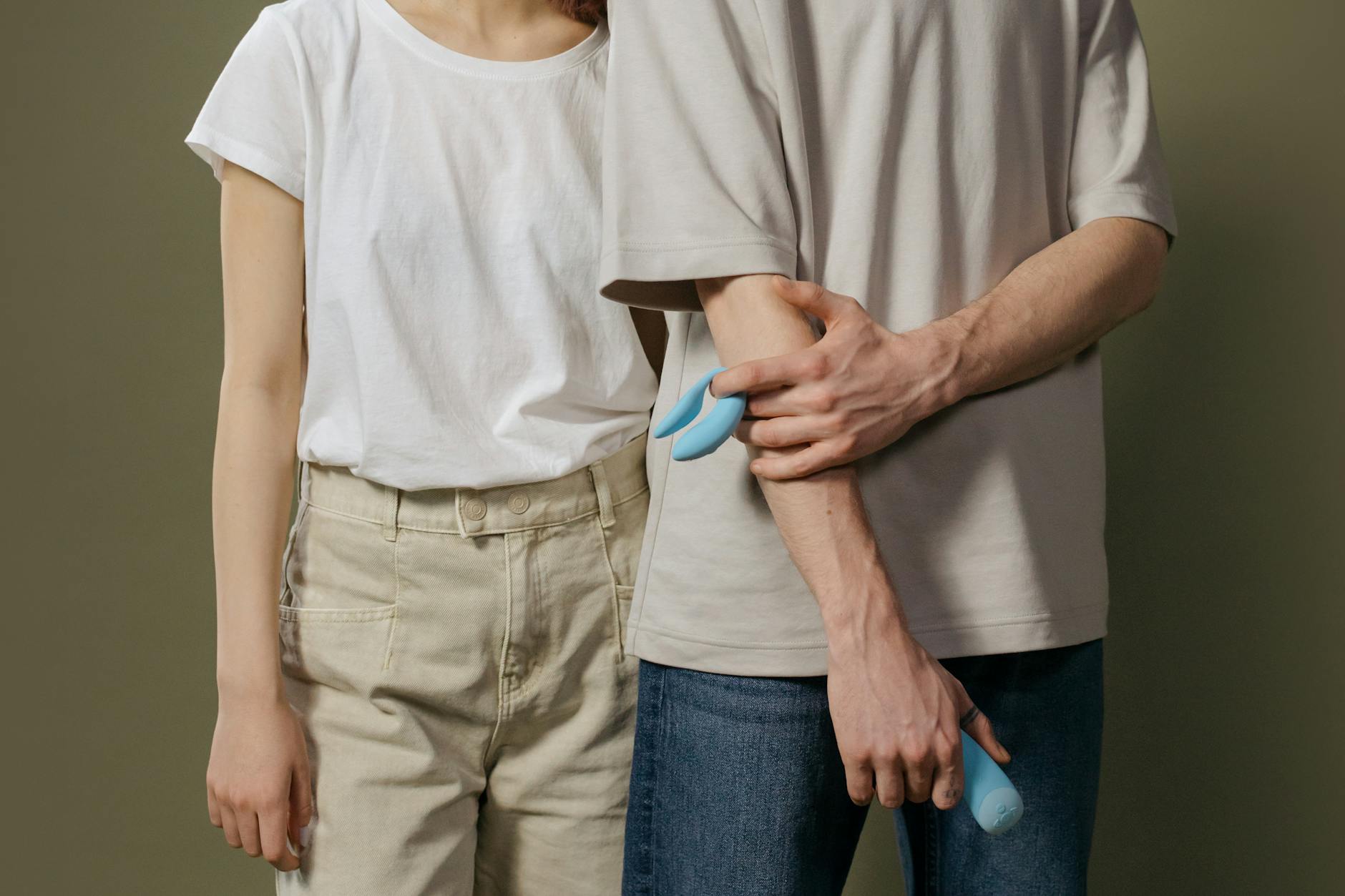 A young couple standing together indoors, holding a blue vibrator, symbolizing modern intimacy and connection across distance.
